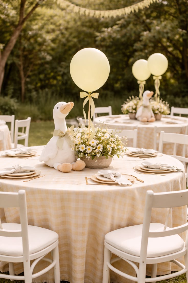 Round lawn tables dressed in yellow and white gingham with daisy centrepieces, plush geese and pastel balloons beneath the trees