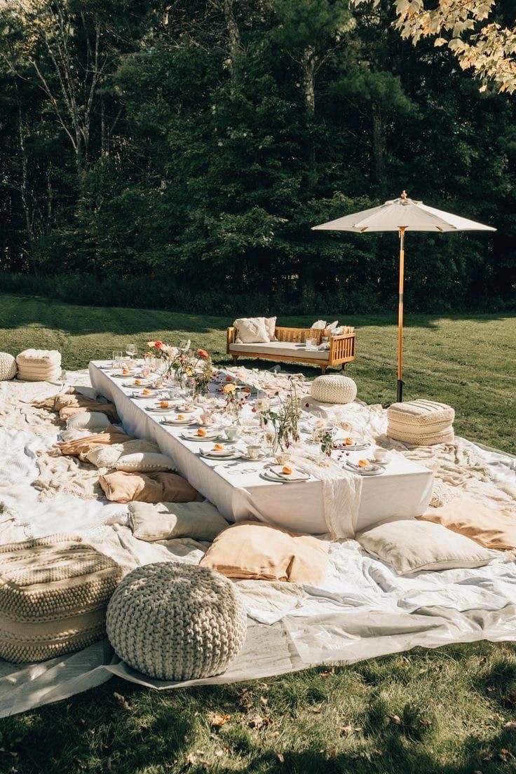 A long lawn table dressed in white linen with glassware, blooms and floor cushions beneath a cream umbrella, trees behind